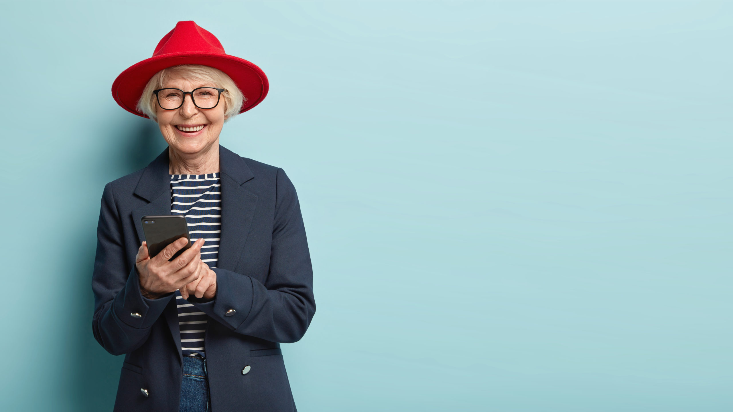 Overjoyed senior lady in stylish apparel and hat chats with friends online, happy to learn using modern gadgets, being advanced user of cell phone, spends leisure time at home, stands indoor alone
